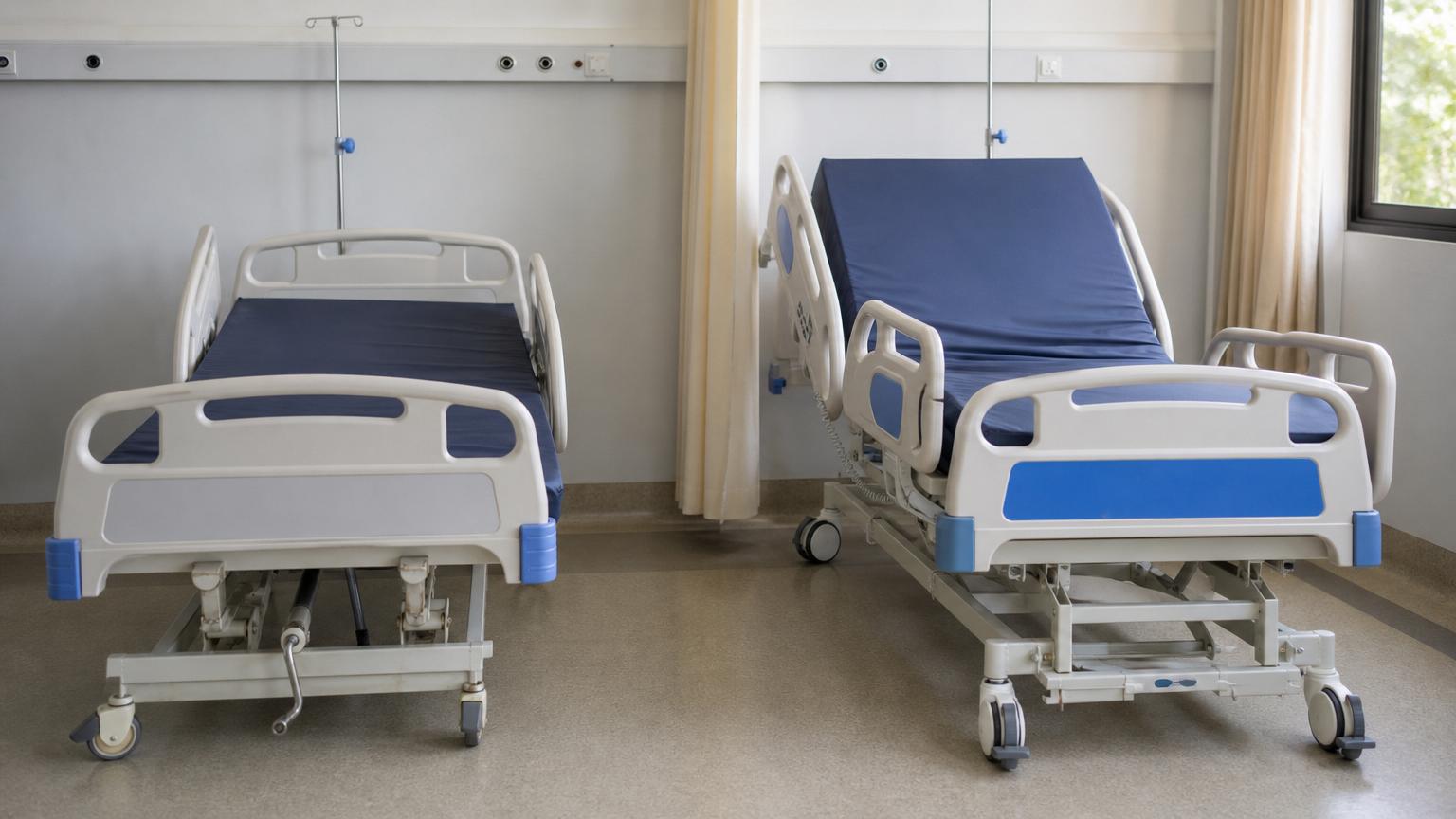 Two adjustable hospital beds with blue mattresses side-by-side in a bright clinic room, one flat and one with the headrest elevated.